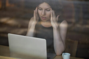 Woman with headache holding temples, sitting in front of laptop.